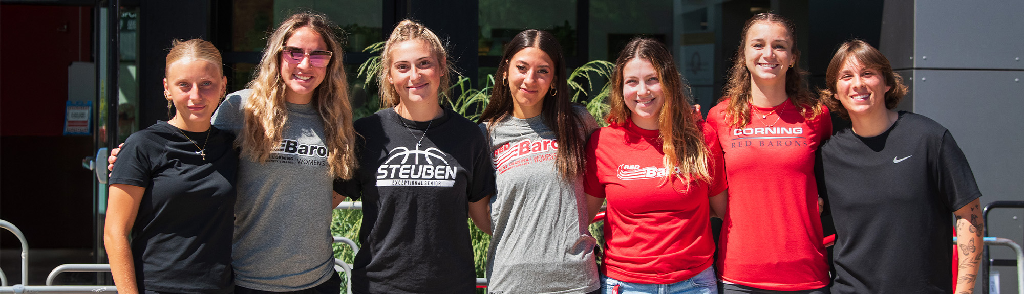 A group of seven people stand side by side outdoors in bright sunlight, with arms around one another. They are wearing casual athletic clothing, including T‑shirts with printed logos such as “Baron,” “Steuben Basketball,” and “Corning Red Barons,” along with shorts or jeans. Behind them is a building with large windows and greenery visible in planters.