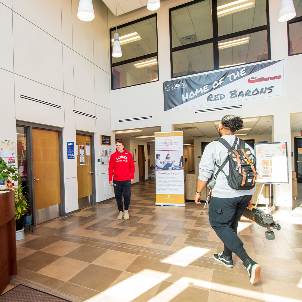Two students in a modern lobby walking by one another. On student is wearing a red sweatshirt and the other is wearing a gray sweatshirt and carrying a skateboard. 