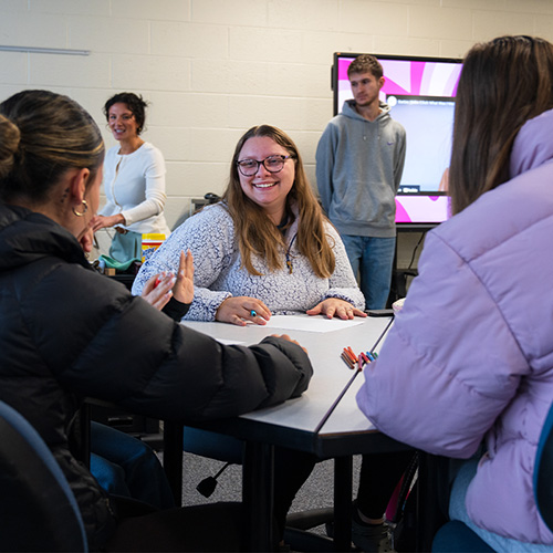 A group of people are seated around a rectangular table in a classroom setting, engaged in discussion. The table has several colored markers on it. In the background, there is a large screen displaying a pink and white graphic, and two individuals are standing near it. The room has white brick walls and a bright, well-lit atmosphere.