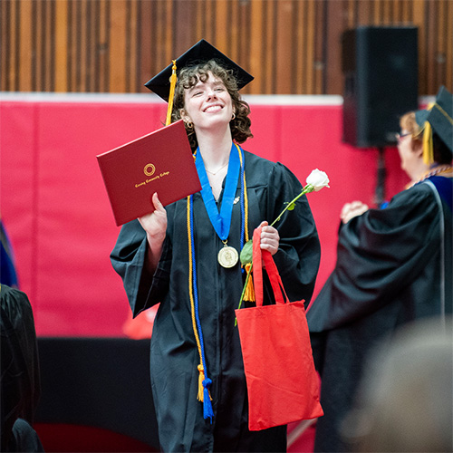 female student dressed in graduation regalia, smiling and holding up her diploma