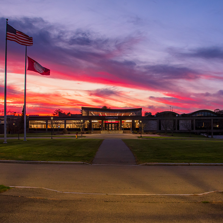 CCC's Commons building in front of a sunset with red, blue and purple skies. American flag and College flag sailing to the left of the building.