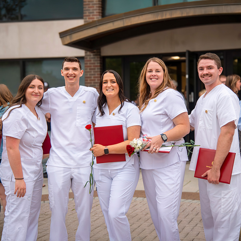 Six people wearing white medical-style uniforms stand together outdoors in front of a brick building entrance. Several people are holding red folders and flowers, and they are positioned close together as if posing for a group photo.