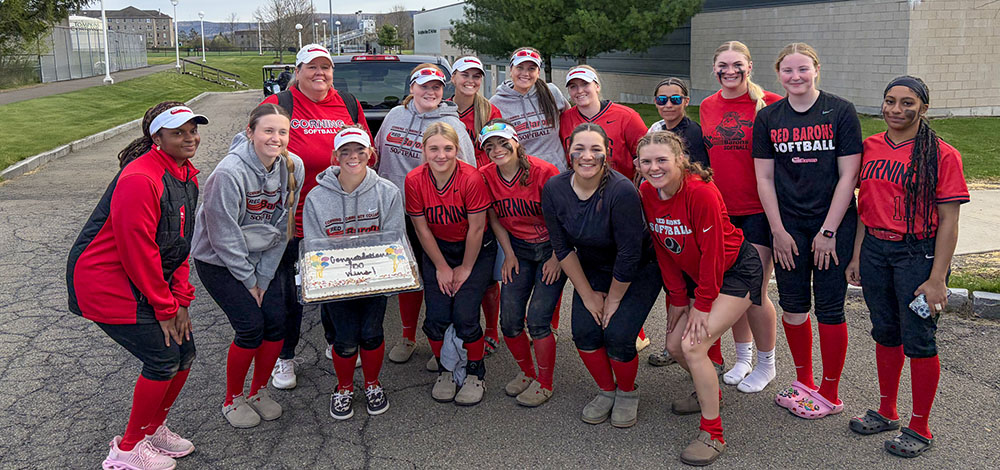 A group of softball players wearing red, black, and gray uniforms pose together outdoors on a paved area near a building and parked vehicles. One person in the front holds a rectangular cake, and several uniforms display the text “Red Barons Softball.”