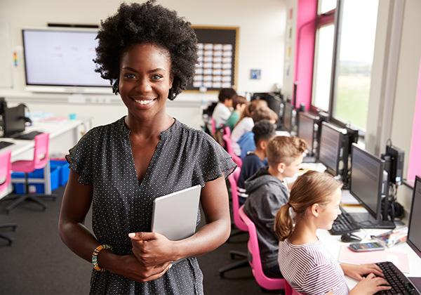female teacher holding a tablet while student work on computers next to her