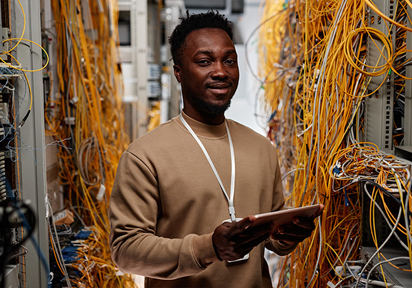 african american man standing with a tablet in a room of servers with yellow exposed wires